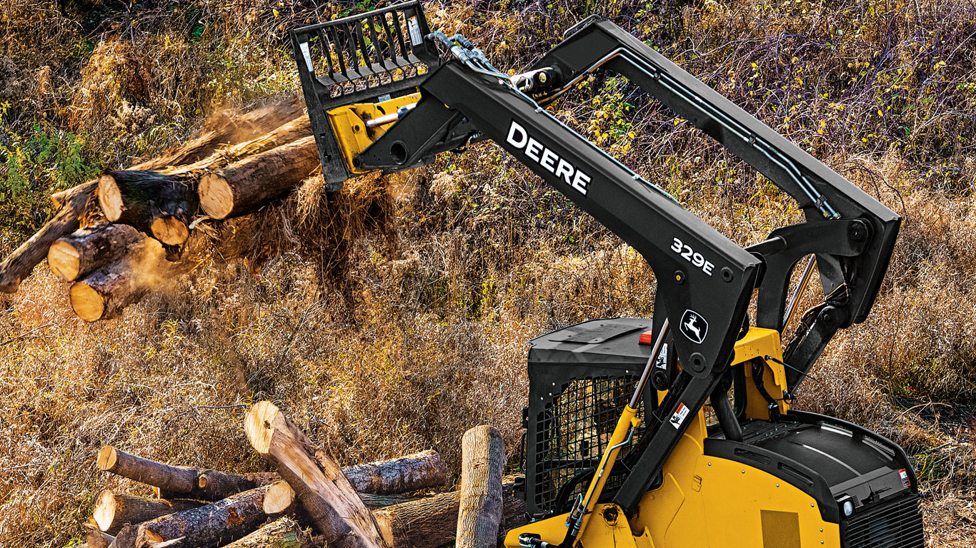 Une chargeuse à direction différentielle John Deere équipée de l’accessoire fourche à palette dépose des grumes.